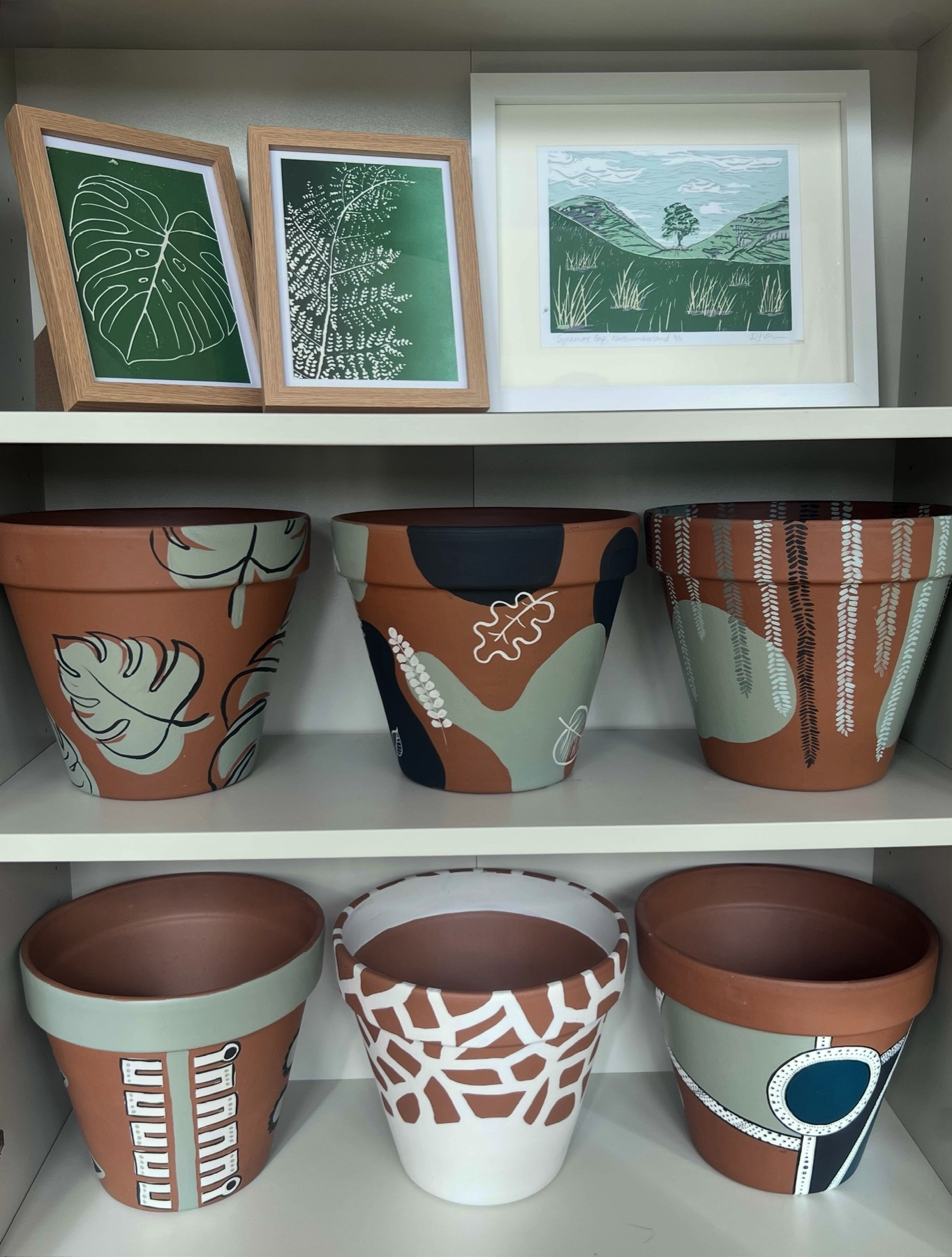A shelf displaying hand-decorated plant pots and framed landscape prints